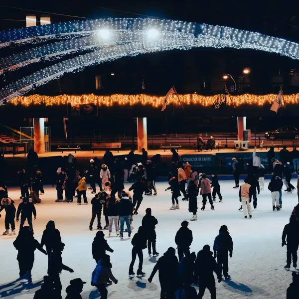 Skaters on a floodlit outdoor ice rink at night under an arch of string lights